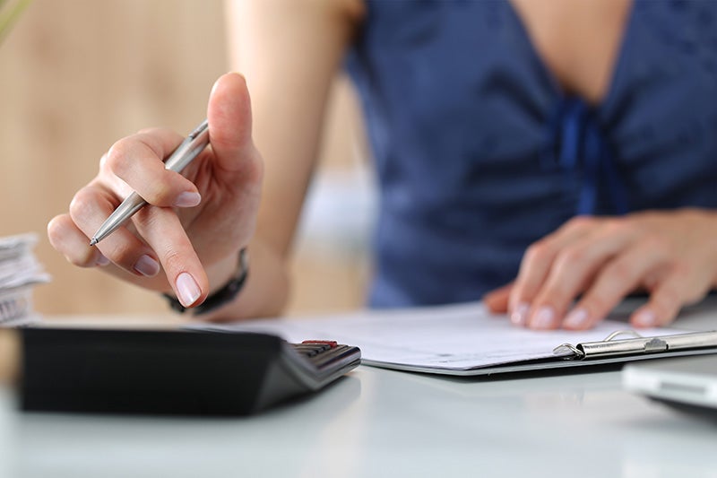 A person's hand with a pen pressing a button on a calculator, with paperwork 